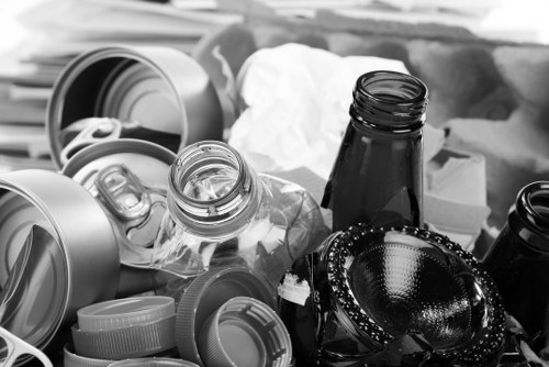 A collection of discarded packaging materials including several crushed aluminum cans, plastic bottles with visible threading and caps, and various opaque plastic containers with screw-top lids, all piled together on a surface, likely part of a waste collection or clearance operation. Some paper or tissue-like material is partially visible among the containers. The objects are arranged casually, with the aluminum cans lying on their sides, the plastic bottles leaning against each other, and the lids scattered on top. The environment appears to be an outdoor or semi-outdoor setting, possibly on a driveway or pavement near a building, indicating rubbish removal activity by Office Clearance Leyton as part of their waste management services in the Leyton area. The overall tone is neutral and descriptive, capturing the physical characteristics and arrangement of the rubbish elements currently being prepared for removal.