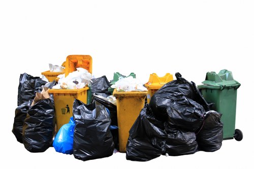 A collection of waste bins and large black rubbish bags arranged outdoors on a paved surface, with a variety of coloured bins including orange, yellow, green, and blue, used for waste collection. The bins are filled with mixed household and general waste, some with lids open or displaced, revealing paper and plastic waste inside. Several black rubbish bags are stacked around the bins, appearing full and tied, with a mix of textured plastic and fabric-like surfaces. The background is plain white, with no additional environmental context visible, highlighting the focus on the waste collection items as part of rubbish removal services, consistent with the offerings of Office Clearance Leyton in East London's postcode area.