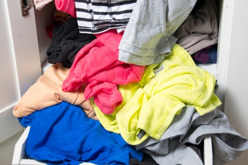 A drawer filled with a variety of crumpled and disorganized clothing items, including a bright pink garment, a yellow shirt, a blue piece of fabric, and several grey and beige textiles, all spilling over the edges of the drawer, which is part of a white storage unit. The surrounding environment appears to be an indoor setting, possibly a bedroom or closet space, with the focus primarily on the cluttered clothing that could require rubbish removal or tidying. The clothing textures are soft and fabric-based, with different colours and materials visible, creating a vivid, casual scene related to laundry or clothing disposal concerns.