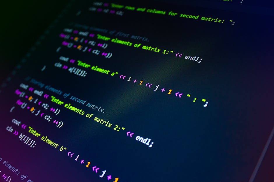 A person dressed in a light blue shirt working at a desk with a laptop displaying colorful lines of programming code on its screen. The individual is holding a pen and appears to be analyzing or editi