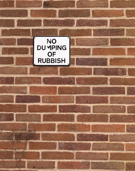 A rectangular metal sign mounted on a red brick wall, which displays the message 'NO DUMPING OF RUBBISH' in black uppercase letters on a white background. The brick wall has a traditional running bond