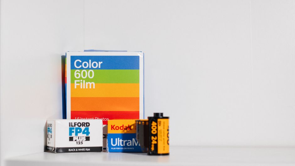 A collection of camera film packs and rolls is arranged on a plain white surface against a light grey background. The items include a multicoloured box labeled 'Color 600 Film' with a blue top and gre