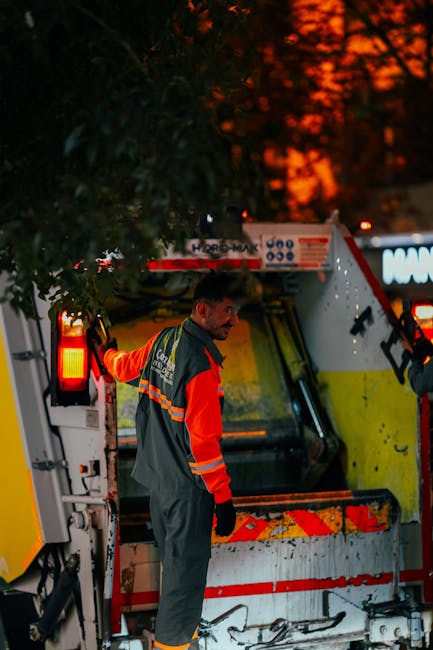 A worker dressed in a green uniform and safety helmet stands on the back step of a waste collection vehicle, which is parked on a suburban street with trees lining the roadside. The rear of the vehicl